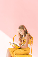 stylish blonde child reading book while sitting on yellow chair on pink