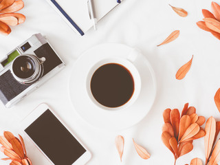 top view of coffee shop concept with coffee cup, notebook, dry leaf, retro camera and smartphone on white fabric background.