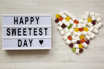 Modern board with text 'Happy Sweetest Day' word and heart made of candies over white wooden background, top view. From above, flat lay, overhead.