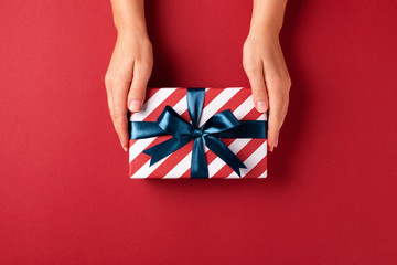 Female's hands holding striped gift box with blue ribbon on red background. Christmas, New Year, Valentine's day and birthday concept.