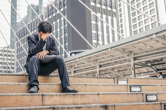 Desperate asian businessman with hands on head sitting on concrete stairs in urban city area. Emotional pain, sadness, business problems and depression concept