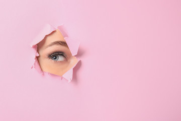 Eye of beautiful young woman visible through hole in pink teared paper - obrazy, fototapety, plakaty