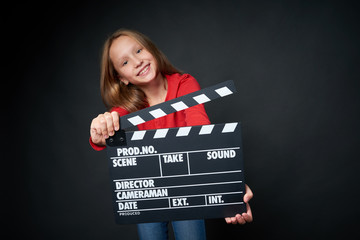 Happy smiling girl holding clap board, over dark background. Shallow depth of field, focus on...