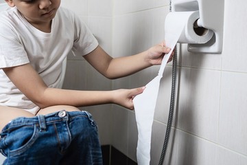 Asian boy sitting on toilet bowl holding tissue paper  - health problem concept