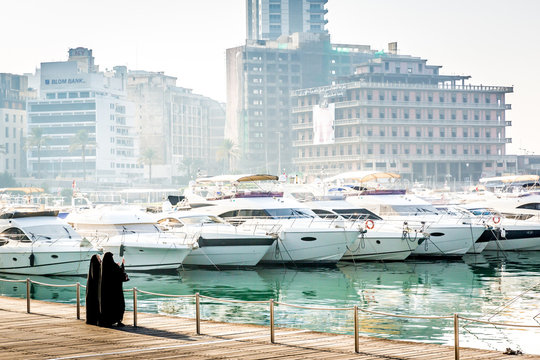 Two woman dressed with black arab traditional clothes in a marine bay full of modern boats with old buildings in the background. Haze air.