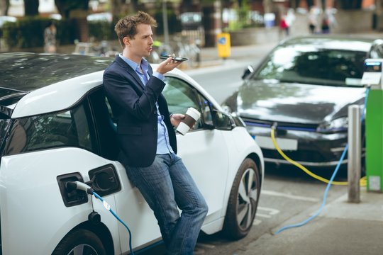 Businessman talking on smartphone while charging electric car