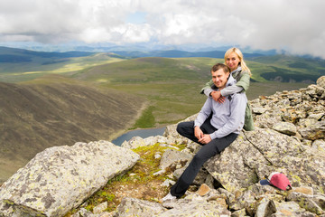 The man sits on a rock, looks into the camera, behind him a blond woman smiles against the background of clouds, blue lake, valley of hills. A loving couple hugs at the top of a mountain in the Altai