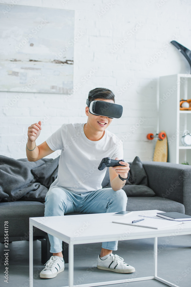 Wall mural excited young man in virtual reality headset playing with joystick at home