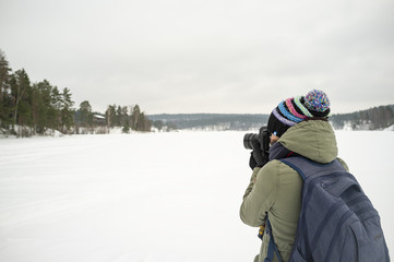 Fototapeta na wymiar A woman photographer in warm clothes holds a camera in her hands and takes pictures of the winter landscape. Against the backdrop of the lake, forest and hut, cold winter, overcast day.