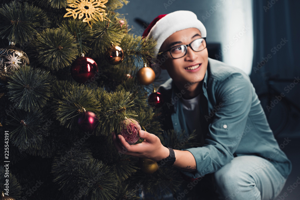 Wall mural young happy asian man in santa hat decorating christmas tree and looking away