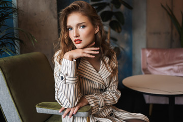Young beautiful woman with wavy hair in striped trench coat leaning on hand thoughtfully looking in camera while spending time in modern cafe