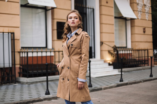 Pretty thoughtful girl in trench coat dreamily looking aside while walking around cozy city street