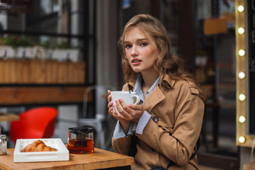 Young attractive woman in trench coat dreamily looking in camera while holding teapot and white cup outdoor at cozy cafe terrace