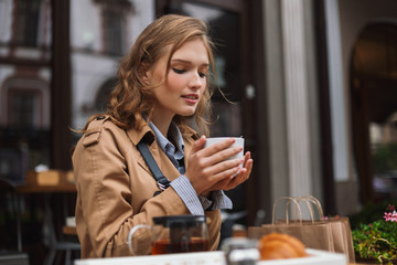 Beautiful girl in trench coat dreamily drinking tea while spending time outdoor at cozy cafe terrace