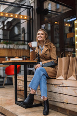 Beautiful smiling girl in trench coat and jeans joyfully looking in camera holding white cup of tea in hand while spending time outdoor at cozy cafe terrace