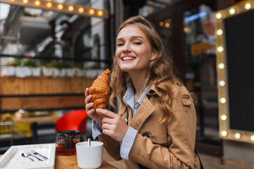 Young smiling woman in trench coat happily looking in camera holding croissant in hand while spending time outdoor at cozy cafe terrace