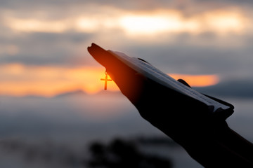Silhouette of human hand holding bible and cross, the background is the sunrise., Concept for...