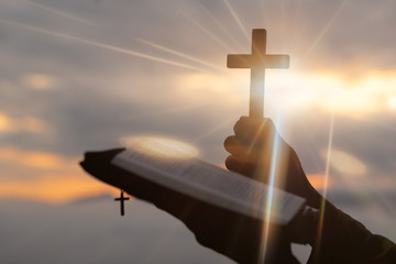 Silhouette of human hand holding the cross with bible, the background is the sunrise., Concept for...