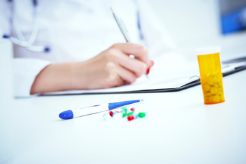 Pills and electronic thermometer laying on the table with female medicine doctor working on background. Medicine doctor prescribes medicine to the patient. Healthcare and medical concept.