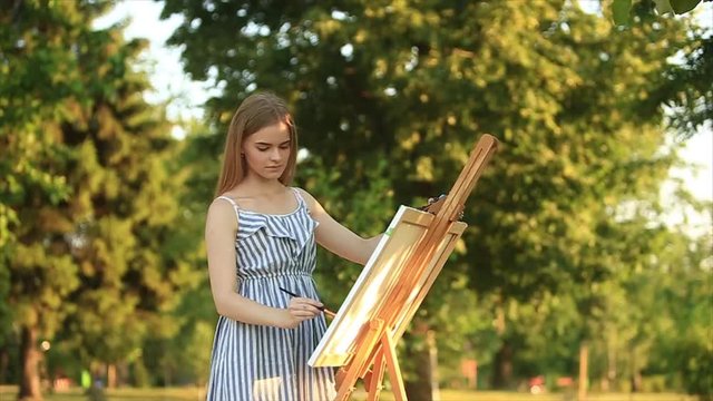 Beautiful girl standing in the park and draws a picture using a palette with paints and a spatula.