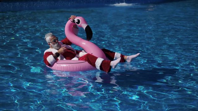 Handsome Santa floating in flamingo inflatable ring into the swimming pool with cocktail glass in hand, chilling and enjoying tropical New Year. Pleasure and happiness concept. Inspiration background