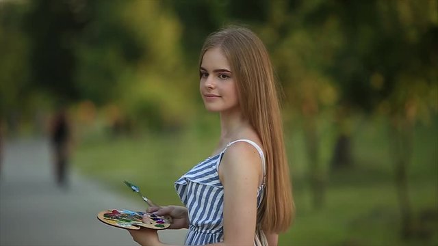 Beautiful girl artist is standing in the park and holding palette with paints