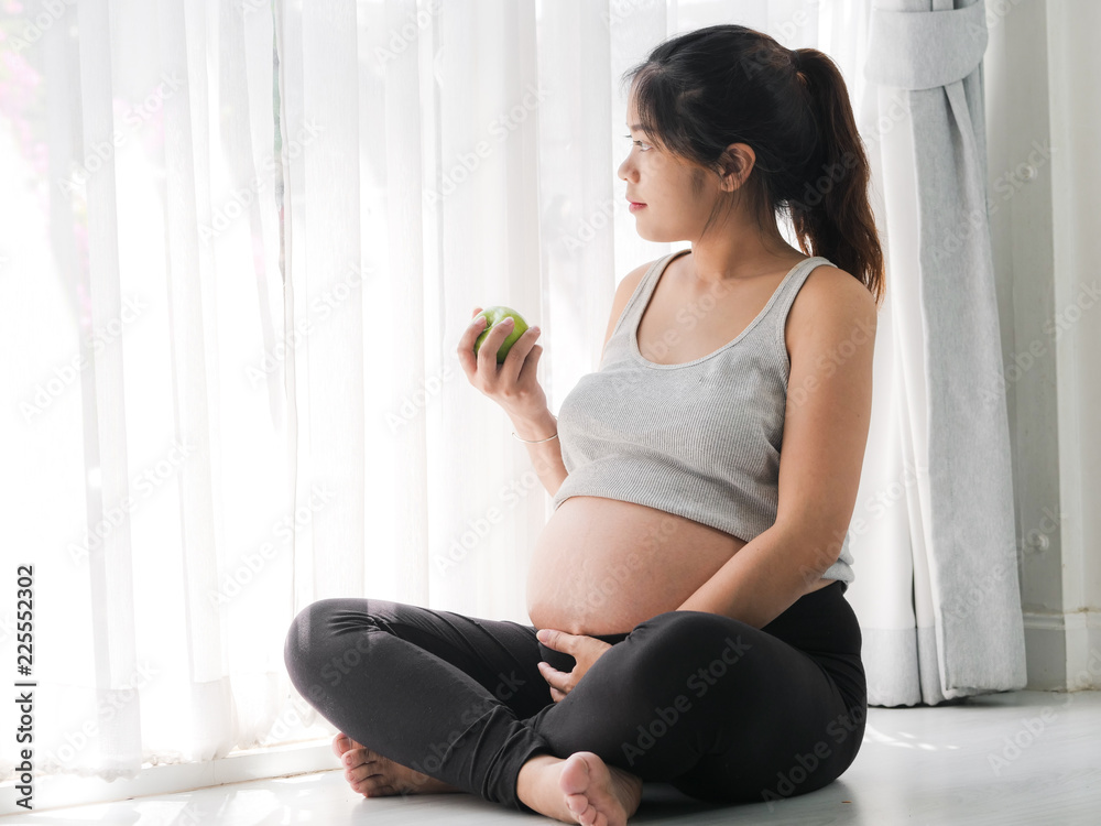 Wall mural happy asian woma holding green apple in her hand and sitting on floor near window, lifestyle concept