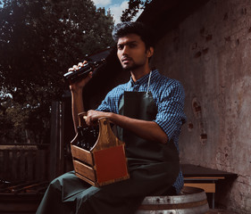 Indian brewer in uniform sitting on a wooden barrel and drink craft beer.