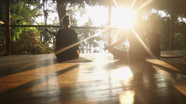 A group of travelers sitting in meditation silhouetted against the evening sun during sunset on a wooden porch overlooking the rainforest and jungle leading out to the beach.