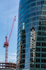 Naklejka premium A red tower crane stands besides a building under construction. The building has reflective glass. It reflections other buildings. The crane can be seen. A blue sky with clouds are in the backgound.