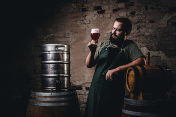 Brewer in apron standing near barrels and drinks craft beer at old brewery factory. - obrazy, fototapety, plakaty