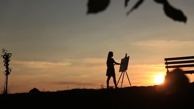 Silhouette of a blonde girl paints a painting on the canvas with the help of paints. A wooden easel keeps the picture. Summer is a sunny day, sunset