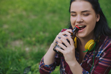 Brunette girl opens a bottle of beer with her teeth. Pretty student opens a beverage using her...