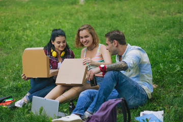 Group of students sit on the grass ans open the boxes with pizza. Pretty girl smiles and hold a box from pizza. Handsome man with a tatto looks into the box.