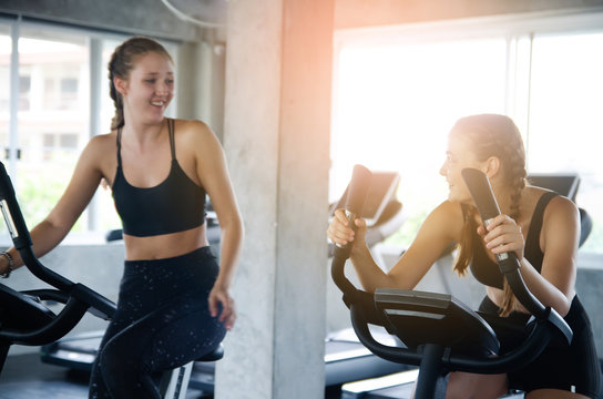 Young People Talking And Smiling While Working Out On Bike At Gym. Friends In A Conversation While Cycling On Stationary Bike In Fitness Centre. Group Of Happy People Working Out At Spinning Class.