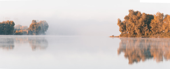Beautiful foggy morning. Wide panorama of autumn lake with fog over  water and reflections of trees.