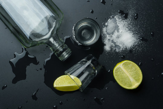top view of an empty bottle of silver tequila with the tequila shot glasses, the lime slices and salt on the wet table