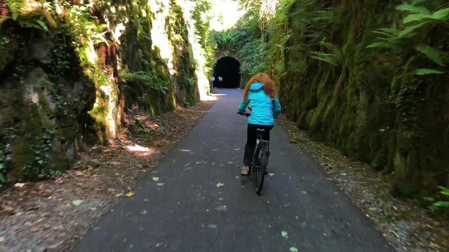 Young redhead woman casually cycling through the beautiful green tunnel covered with plants. Waterford Greenway.