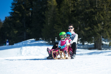 Mother and children having fun on sledge with mountain scenery in background. Active mom and kids with safety helmets. Winter sport for family. Little kids outside, swiss Alps, healthy lifestyle 