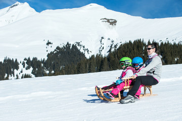 Mother and children having fun on sledge with mountain scenery in background. Active mom and kids with safety helmets. Winter sport for family. Little kids outside, swiss Alps, healthy lifestyle 