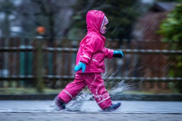 Girl is having fun in water on street in cold autumn day, girls splashing water in rain, happy and cheerful girl enjoying cold weather, kid in pink rain coats and rubber boots, running in rain