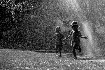 Girls are having fun in water in park, hot summer in garden, girls running in water drops, happy and cheerful girls enjoying cold water in hot summer day, people black and white silhouettes 