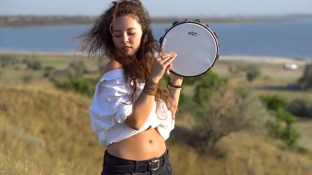 Curly Gipsy brunette girl playing on tambourine in the field on the slope of Gulf