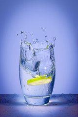 Lemon slice falls on mineral water and splashing water on blue background