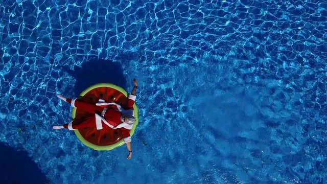 Aerial view: Santa lie on the watermelon inflatable ring and calling to come makes a hand gesture. Concept of satisfaction and xmas vacation. Cheerful man celebrate life, summertime and happiness