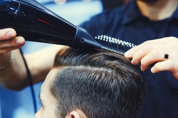 Male barber makes hair styling of a young man using a dryer