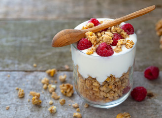 White yogurt with muesli in glass bowl with pieces raspberries on top on natural wooden desk.