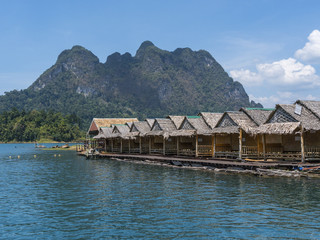 Naklejka premium Wooden Thai traditional floating houses on a lake with mountains and rain forest in the background during a sunny day at Ratchaprapha Dam at Khao Sok National Park, Thailand