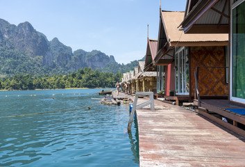 Fototapeta premium Wooden Thai traditional floating houses on a lake with mountains and rain forest in the background during a sunny day at Ratchaprapha Dam at Khao Sok National Park, Thailand