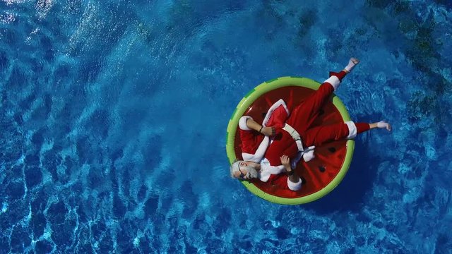 Aerial view: Santa lie on the watermelon inflatable ring and calling to come makes a hand gesture. Concept of satisfaction and xmas vacation. Cheerful man celebrate life, summertime and happiness